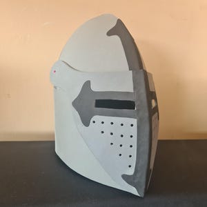 May include: A light gray medieval helmet with dark gray accents. The helmet features a visor with small circular perforations and a cross-shaped design. The helmet is made of a sturdy material and is designed to protect the head.