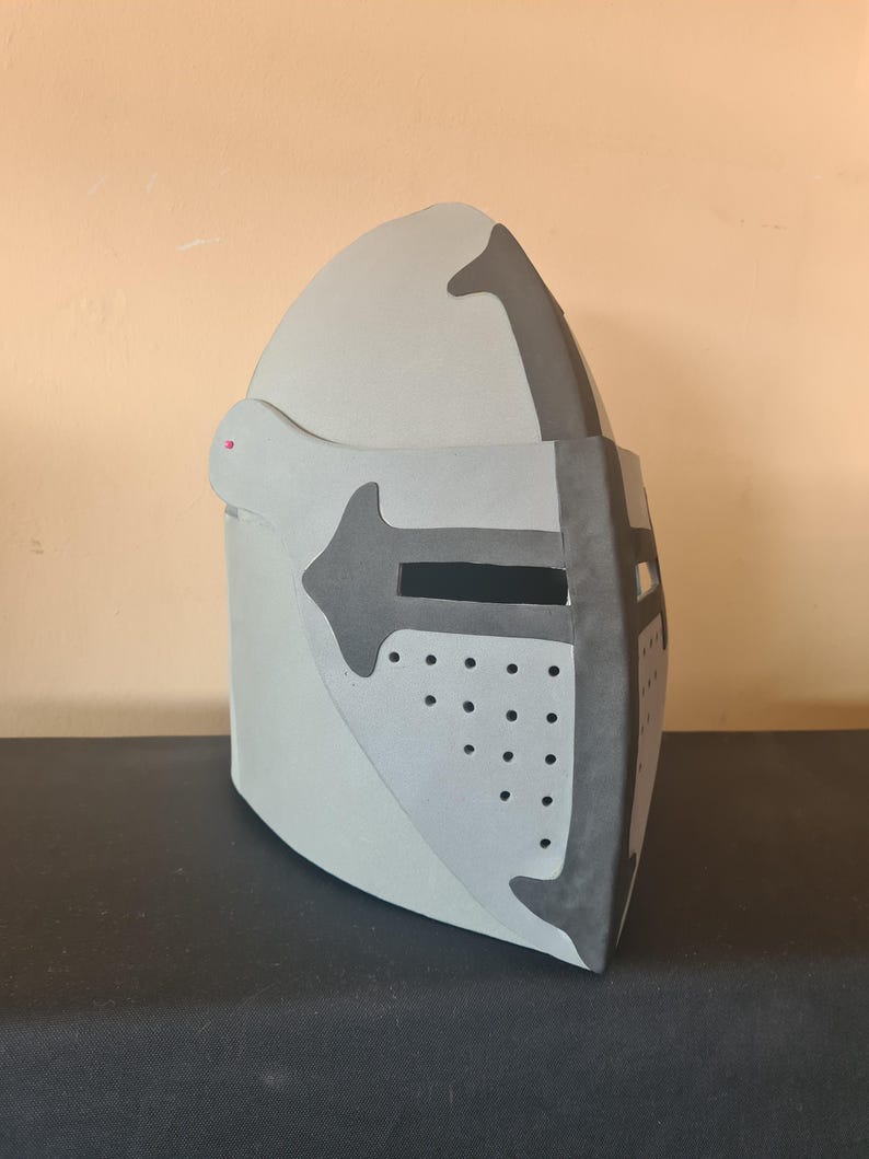 May include: A light gray medieval helmet with dark gray accents. The helmet features a visor with small circular perforations and a cross-shaped design. The helmet is made of a sturdy material and is designed to protect the head.