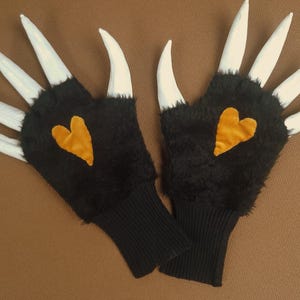 May include: A pair of black gloves with white claw-like extensions. Each glove features a gold heart-shaped patch. The gloves have a black ribbed cuff and are displayed on a brown surface.