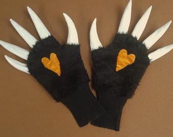 May include: A pair of black gloves with white claw-like extensions. Each glove features a gold heart-shaped patch. The gloves have a black ribbed cuff and are displayed on a brown surface.