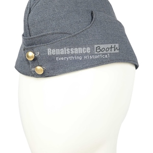 May include: A gray military-style hat with a curved brim and two gold-colored buttons. The hat is displayed on a white mannequin head. The text "Renaissance Booth Everything Historical" is printed on the hat.