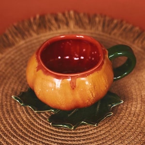 May include: A ceramic pumpkin-shaped mug with a green handle and a dark red interior. The mug sits on a green leaf-shaped saucer. The exterior of the mug is orange with brown accents.