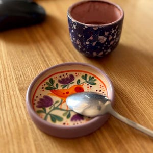 May include: A small, dark blue ceramic cup with white speckles sits next to a round, light purple ceramic saucer. The saucer features a hand-painted design of an orange bird surrounded by purple flowers and green leaves, with a silver spoon resting on top.