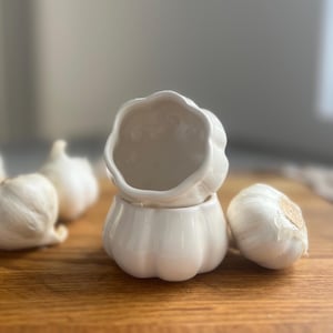 May include: A white ceramic garlic keeper, shaped like a garlic clove, with a lid, on a wooden surface. Several whole garlic bulbs are scattered around the keeper. The keeper is designed for kitchen use.