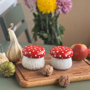 May include: Two ceramic mushroom-shaped containers with glossy red caps featuring white polka dots and textured off-white stems. The containers are placed on a wooden cutting board alongside a gourd, a small green pumpkin, two walnuts, and an apple.