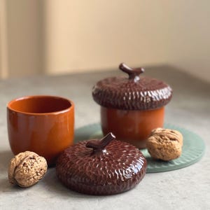 Op de afbeelding: Een opstelling van keramische eikelvormige containers en een kleine oranje beker. De eikelcontainers zijn bruin met getextureerde deksels en een steelachtig handvat. Een walnoot ligt naast de containers op een lichtgroene plaat.