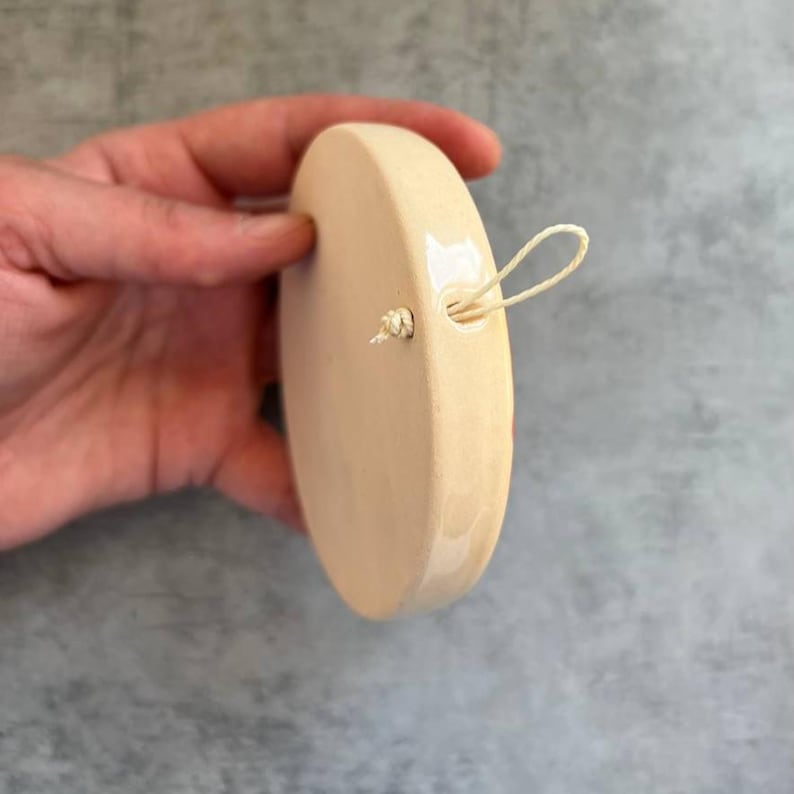 May include: A circular, cream-colored ceramic ornament with a smooth, glossy finish. The object features a small hole near the top edge, through which a thin, light-colored string is looped and knotted, allowing for easy hanging display.