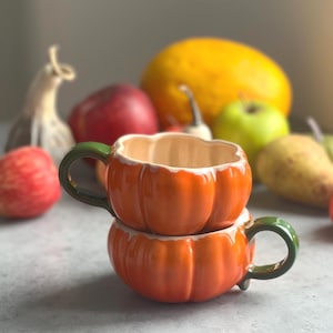 May include: Two stacked ceramic mugs shaped like orange pumpkins with glossy finishes and dark green handles. The mugs feature vertical ridges mimicking the texture of a pumpkin, with a cream-colored interior and a smooth, rounded rim design.