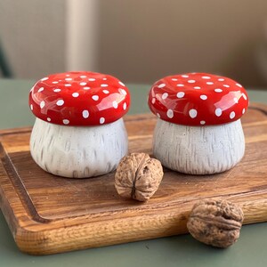 May include: Two ceramic mushroom-shaped containers with glossy red caps featuring white polka dots and textured off-white stems. Two whole walnuts are positioned in front of the containers on a wooden board with a visible grain and grooved edges.