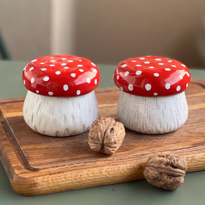 May include: Two ceramic mushroom-shaped containers with glossy red caps featuring white polka dots and textured off-white stems. Two whole walnuts are positioned in front of the containers on a wooden board with a visible grain and grooved edges.