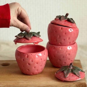 May include: Pink ceramic strawberry-shaped containers with lids. The containers have a textured surface with small indentations, resembling strawberry seeds. The lids are topped with green and brown leaf details. One lid is being lifted, revealing the inside of the container.