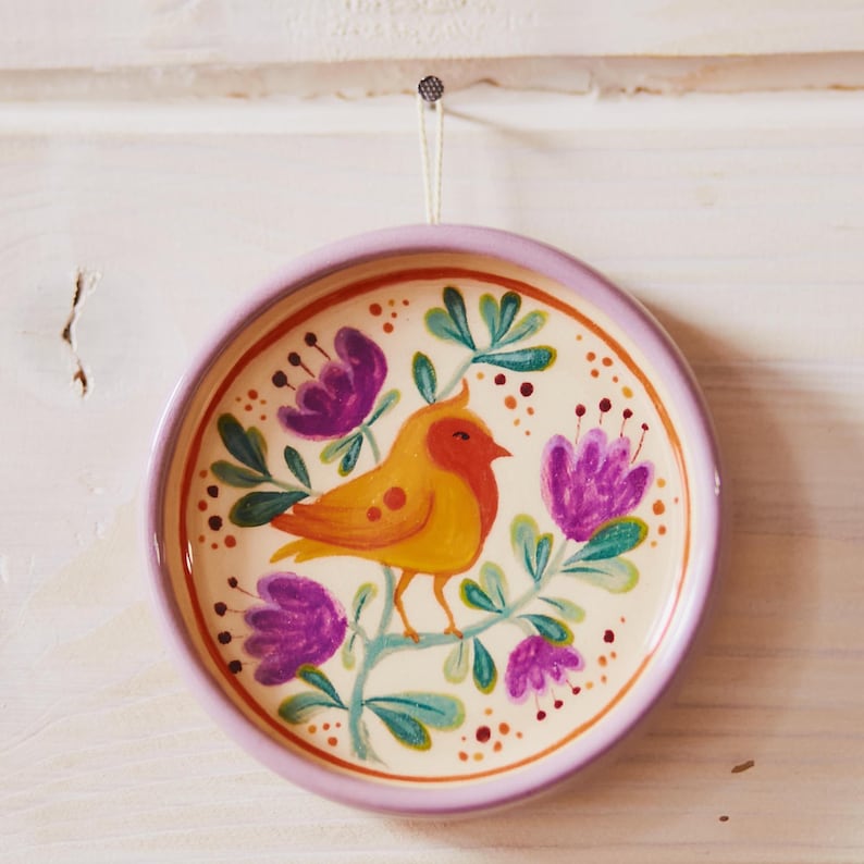 May include: A round ceramic wall plate with a light lavender rim, featuring a hand-painted design of a yellow-orange bird perched on a branch with purple flowers and green leaves, suspended by a thin white string from a small metal hook.
