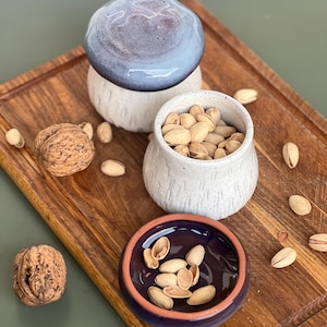 May include: A wooden serving board displays ceramic bowls and nuts. One bowl is filled with pistachios, another with pistachio shells, and a third with a blue and white lid. Walnuts and pistachios are scattered around the bowls.