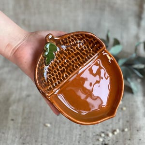 May include: A ceramic acorn-shaped dish with a glossy, brown finish. The top half has a textured pattern, while the bottom half is smooth. A small green leaf detail adds a pop of color. This decorative dish is ideal for serving snacks or as a unique decor piece.