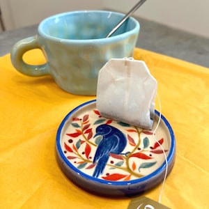May include: A light blue ceramic mug with a textured surface and a silver spoon inside, paired with a round ceramic tea bag rest featuring a painted blue bird perched on a branch with red and orange leaves, holding a white tea bag.
