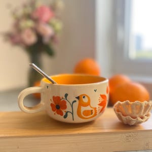 Puede incluir: Una taza de cerámica color crema con interior naranja, con un diseño pintado a mano de pájaros y flores en naranja y negro. Una pequeña cuchara plateada descansa dentro. Naranjas y una galleta en una tabla de madera.