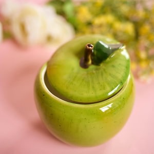 May include: A ceramic apple-shaped container with a lid. The container and lid are a glossy green with a brown stem and a green leaf. The container is on a pink surface with blurred flowers in the background.