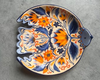 May include: A ceramic dish shaped like a stylized bird with a cream-colored base. The surface features hand-painted floral patterns in vibrant orange and deep blue, with a small blue head and eye detail on the right side and scalloped tail edges.