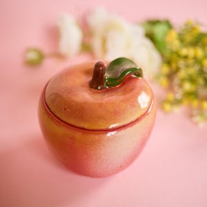 May include: A ceramic apple-shaped trinket box with a lid. The box is primarily peach-colored with red accents, and a green leaf and brown stem on top. The box is on a pink surface.