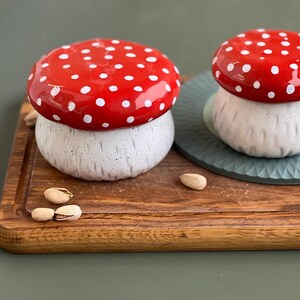 May include: Two ceramic mushroom-shaped containers with glossy red caps featuring white polka dots and speckled white stems. The stems have a textured, vertical grooved finish. The containers are placed on a wooden board with scattered pistachios.