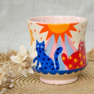 May include: A ceramic mug with a cream-coloured base featuring a hand-painted design of a blue cat with dark blue spots and a red cat with yellow flower patterns. Above the cats is a bright orange sun with rays, and the base has a blue scalloped trim.