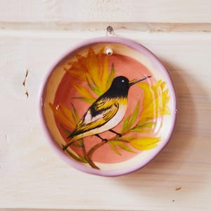 May include: A round ceramic dish with a light purple rim and a hand-painted illustration of a black, white, and yellow bird perched on a branch. The background inside the dish is a soft pink, featuring yellow floral accents surrounding the bird.