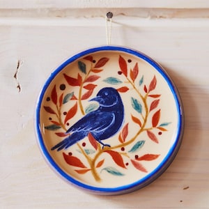 May include: A round ceramic wall plate featuring a hand-painted dark blue bird perched on a branch. The design includes orange, red, and teal leaves surrounding the bird against a cream background, with a solid blue rim and a hanging string.
