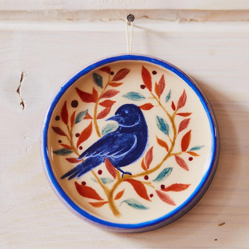 May include: A round ceramic wall plate featuring a hand-painted dark blue bird perched on a branch. The design includes orange, red, and teal leaves surrounding the bird against a cream background, with a solid blue rim and a hanging string.