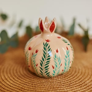 Puede incluir: Figura de cerámica con forma de granada, de color melocotón claro. Decorada con diseños de hojas verdes y flores rojas. La parte superior de la granada tiene forma de corona con detalles rojos. Colocada sobre una superficie tejida. Mide unos 10 cm de alto.