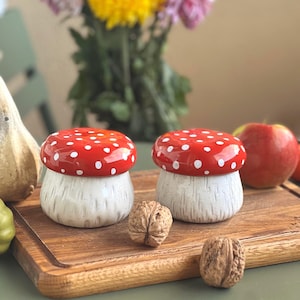 May include: Two ceramic mushroom-shaped containers. The red tops have white polka dots, and the stems are white with a speckled texture. They sit on a wooden board with walnuts and an apple.