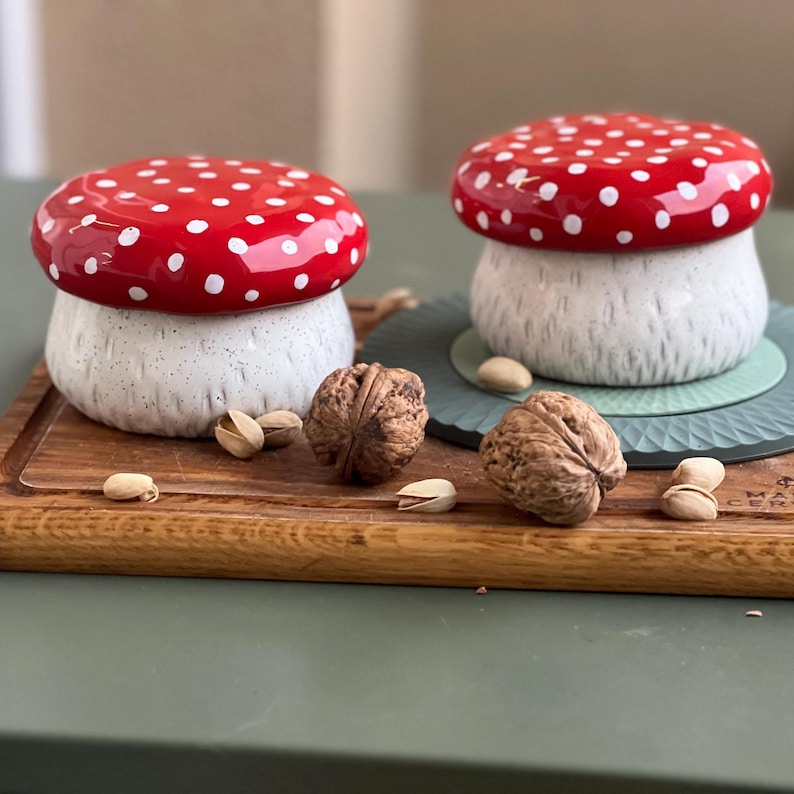 May include: Two ceramic mushroom-shaped lidded jars featuring glossy red caps with white polka dots and textured, speckled off-white stems. The containers are arranged with whole walnuts and pistachio shells on a wooden board surface.