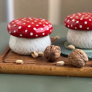 May include: Two ceramic mushroom-shaped lidded jars with glossy red caps featuring white polka dots and textured, off-white stems. The jars are arranged alongside two whole walnuts and several scattered pistachio shells on a wooden surface.