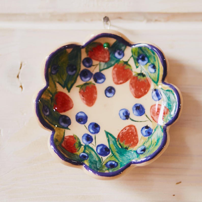 May include: A flower-shaped ceramic dish with a cream-colored base, featuring a hand-painted pattern of red strawberries, blue blueberries, and green leaves. The scalloped edges of the dish are outlined in a deep purple-blue border.