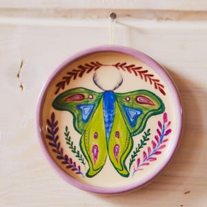 May include: A round ceramic dish with a lavender rim and a cream-colored center. The dish features a hand-painted moth with bright green, blue, and pink wings, surrounded by a decorative border of red and purple leafy sprigs.