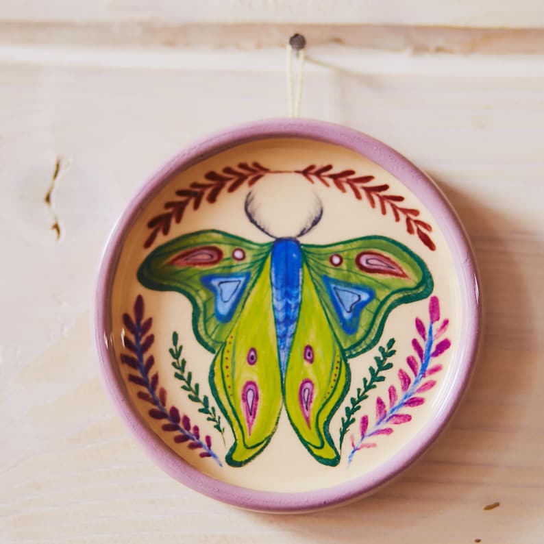 May include: A round ceramic dish with a lavender rim and a cream-colored center. The dish features a hand-painted moth with bright green, blue, and pink wings, surrounded by a decorative border of red and purple leafy sprigs.