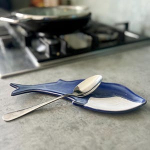 May include: A dark blue ceramic spoon rest shaped like a whale with a white underside detail. A polished stainless steel spoon rests diagonally across the center of the whale-shaped dish, which features a defined tail and fin.