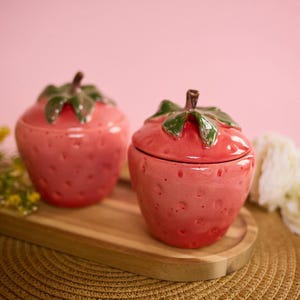 May include: Two ceramic strawberry-shaped containers with lids, in a vibrant pink hue. The lids feature green leaves and brown stems. The containers sit on a wooden tray, ideal for kitchen decor.