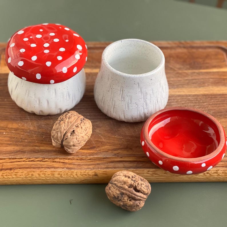 May include: A ceramic mushroom-shaped container with a glossy red cap featuring white polka dots and a textured white base. Beside it sits an open white ceramic cup with a matching red lid featuring white dots, accompanied by two whole walnuts.