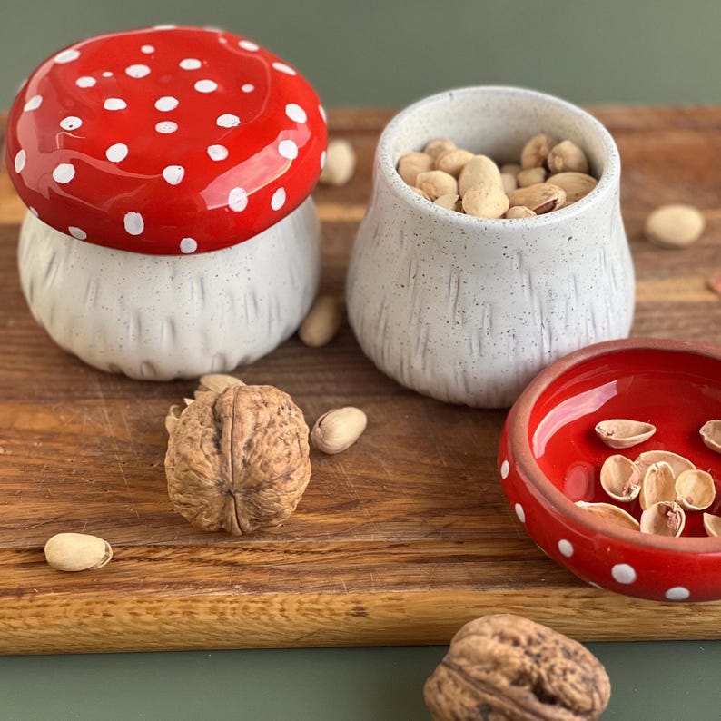 May include: A ceramic mushroom-shaped lidded jar with a red cap and white spots, a matching white speckled ceramic bowl filled with pistachios, and a small red bowl with white dots containing pistachio shells, accompanied by whole walnuts.