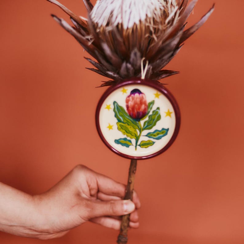 May include: A round, dark red framed ornament featuring a painted illustration of a pink protea flower with green leaves and yellow stars on a cream background, suspended from a stem of a large, dried, spiky brown protea flower.
