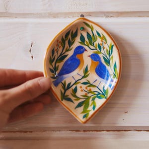 May include: A leaf-shaped ceramic dish featuring a hand-painted design of two blue birds with orange chests perched among green leafy branches and small yellow berries. The dish has a cream-colored background and a brown rim.