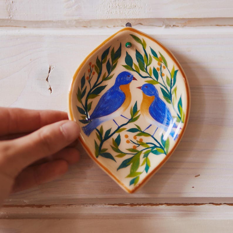 May include: A leaf-shaped ceramic dish featuring a hand-painted design of two blue birds with orange chests perched among green leafy branches and small yellow berries. The dish has a cream-colored background and a brown rim.