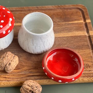 May include: A ceramic mushroom-shaped container with a red cap featuring white spots and a white speckled base, a matching white speckled cup, and a small red dish with white spots, accompanied by two whole walnuts on a wooden surface.