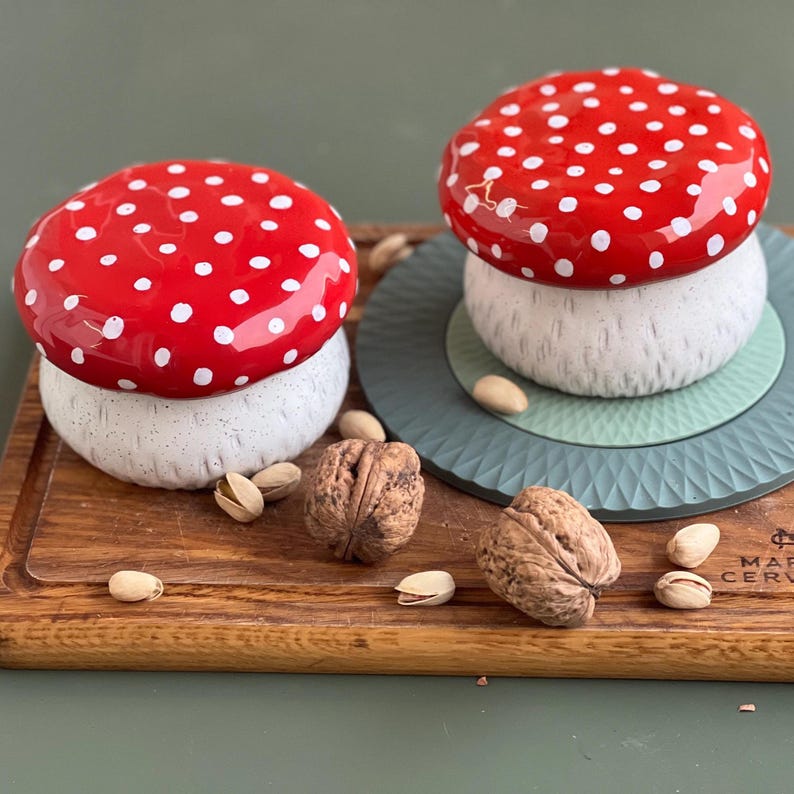 May include: Two ceramic mushroom-shaped containers with glossy red caps featuring white polka dots and textured off-white stems. The mushrooms are arranged with scattered whole walnuts and pistachio shells on a wooden board surface.