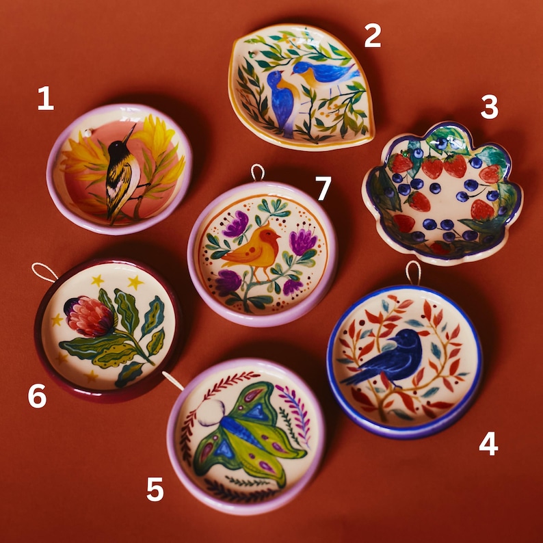 May include: Seven small, round and leaf-shaped ceramic dishes featuring hand-painted designs. The collection includes illustrations of birds, a moth, strawberries, blueberries, and a flower, each numbered one through seven for identification.