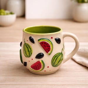 Pode incluir: Caneca de cerâmica creme com interior e alça verdes. A caneca é decorada com fatias de melancia em relevo, melancias inteiras e sementes pretas. Acentos vermelhos e verdes são espalhados pela superfície, criando um tema de verão.