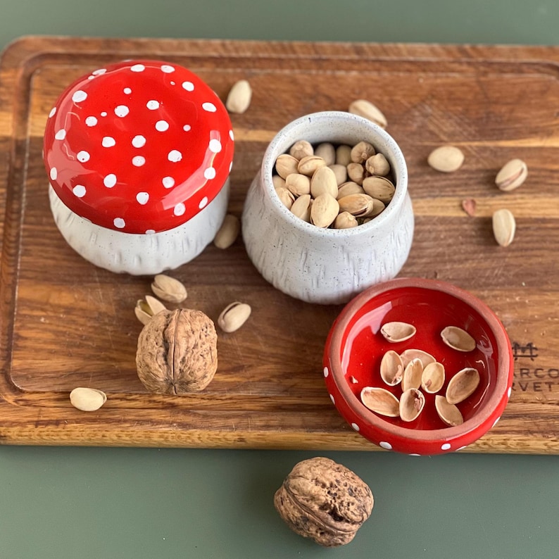 May include: A ceramic mushroom-shaped container with a red cap featuring white polka dots and a textured white base. Beside it sits an open white ceramic jar filled with pistachios and a small red bowl containing empty pistachio shells.