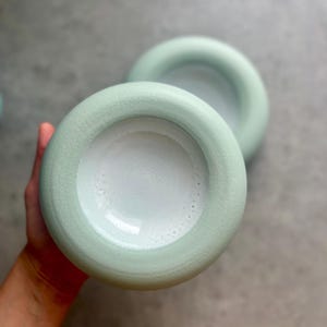 May include: Two pale green ceramic bowls with a rounded rim and a white interior. The bowls are placed on a grey surface. The bowls are similar in size and shape.