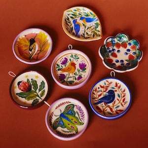 Op de afbeelding: Een verzameling van zeven kleine, ronde en bladvormige keramische schaaltjes met handgeschilderde ontwerpen. De motieven omvatten diverse vogels, een mot, een bloem en aardbeien. Elk schaaltje heeft een gladde glazuurlaag en een ophanglus.