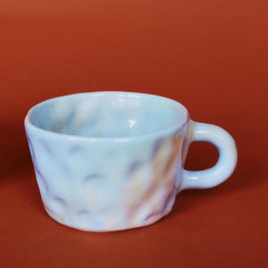 May include: A small, light blue ceramic mug with a textured surface and a rounded handle. The mug has a unique, handmade appearance, with subtle color variations of yellow and orange. The mug is designed for beverages.